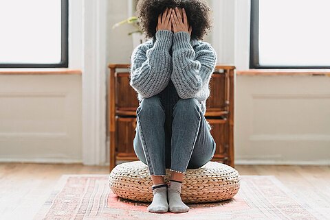 Depressive Frau mit Händen vor dem Gesicht, sitzt auf einem Sitzkissen.