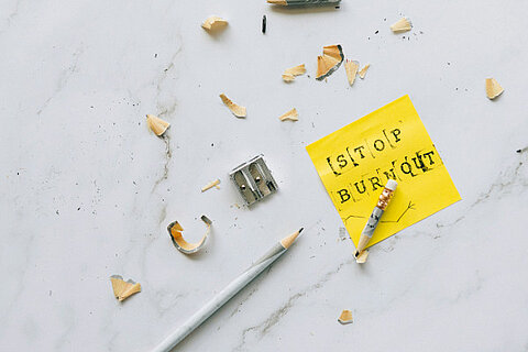Ein gelbes Haftnotiz-Zettel mit der Aufschrift „Stop Burnout" liegt auf einer Marmoroberfläche, umgeben von einem Bleistift, einem Anspitzer und Bleistiftspänen.